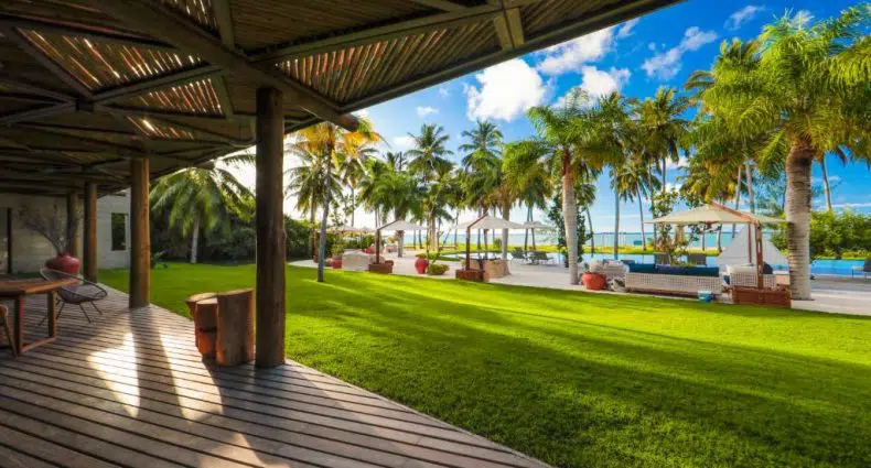 Deck de madeira e pátio coberto com vista para um gramado verde, palmeiras e uma área de piscina com espreguiçadeiras sob um céu azul.