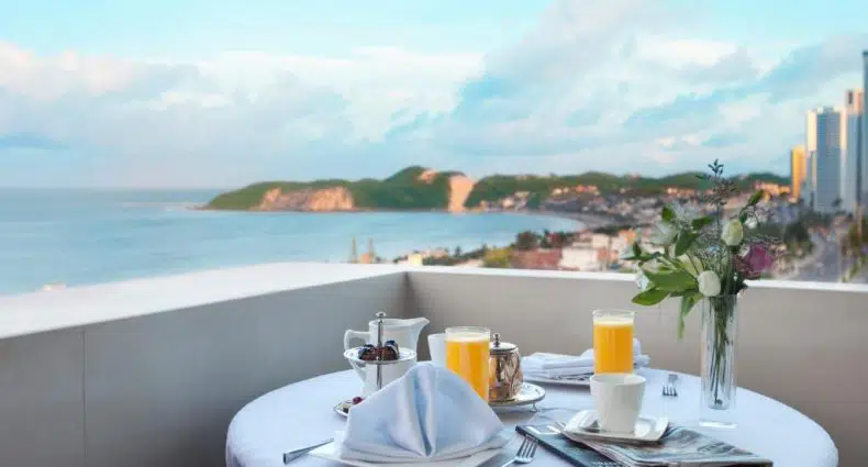 Uma mesa redonda posta para o café da manhã com suco, café e flores em uma varanda com vista para o oceano e a paisagem urbana.