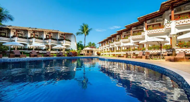Piscina externa com água azul clara, cercada por espreguiçadeiras e prédios do resort sob um céu azul brilhante.