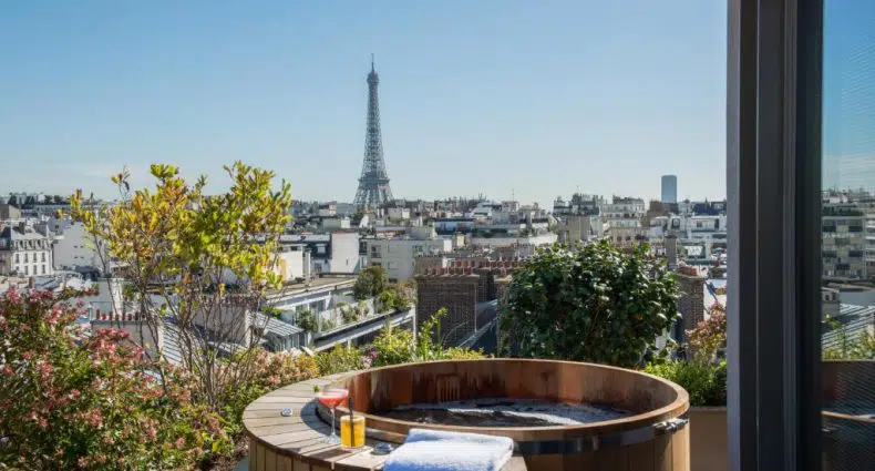Uma banheira de hidromassagem na cobertura do Brach Paris tem vista para os telhados de Paris e para a Torre Eiffel à distância, sob um céu azul claro.