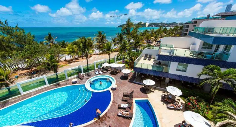 Um hotel à beira-mar com piscinas externas, espreguiçadeiras e palmeiras sob um céu azul brilhante com nuvens dispersas.