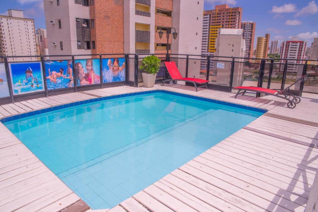 Piscina na cobertura com duas espreguiçadeiras vermelhas, um vaso de plantas e prédios altos ao fundo sob um céu azul.