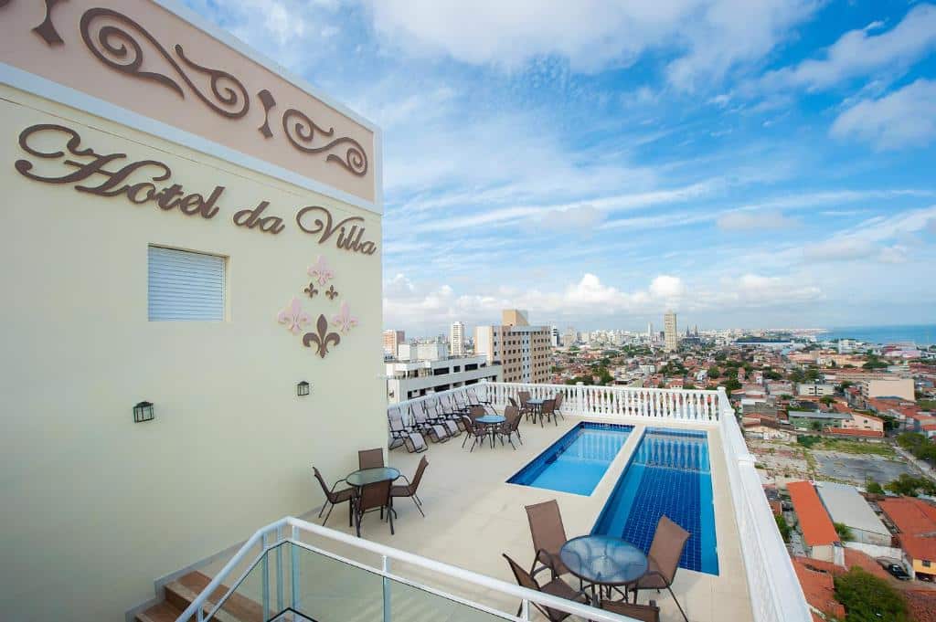 Área da piscina na cobertura com mesas e cadeiras no Hotel da Villa, com vista para a paisagem urbana e a costa sob um céu parcialmente nublado.