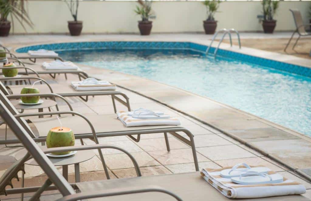 Espreguiçadeiras com toalhas e cocos estão alinhadas ao lado de uma piscina externa de águas claras.