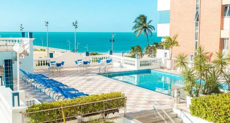 Área da piscina externa com espreguiçadeiras azuis, mesas e vista para o mar ao lado de um edifício moderno em um dia ensolarado.