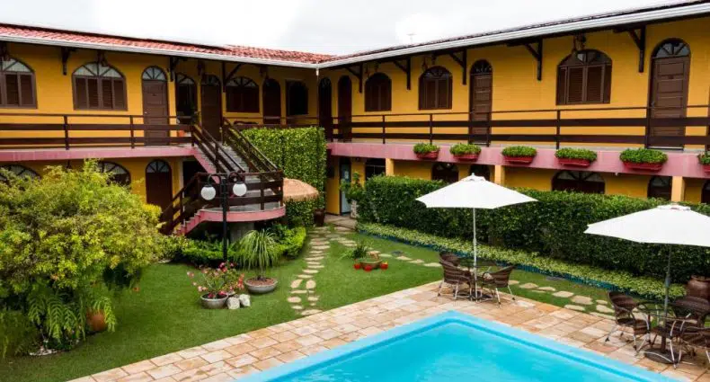 Um pátio com piscina, mesas e guarda-sóis, cercado por um prédio amarelo de dois andares com grades de madeira.