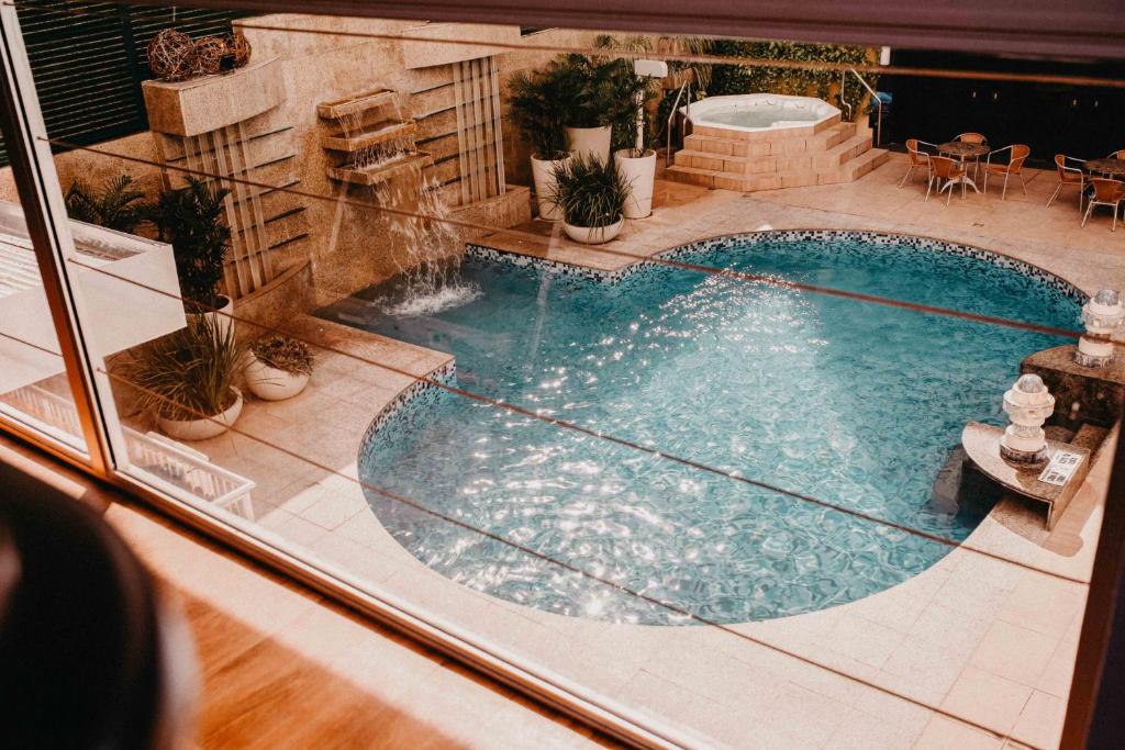 Vista de uma moderna piscina externa com cascata, vasos de plantas, banheira de hidromassagem e cadeiras no pátio ao fundo.