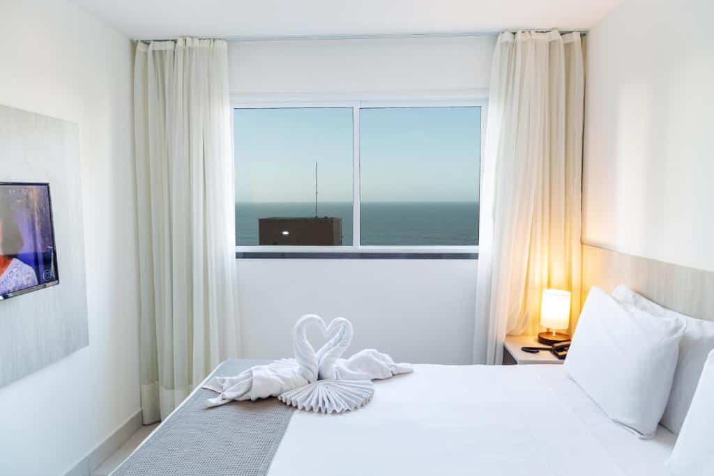 Uma cama bem arrumada com cisnes de toalha, um abajur aceso e uma janela com vista para o oceano em um quarto claro e minimalista.