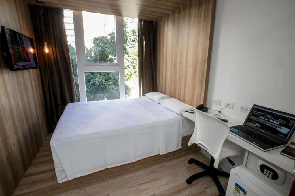 Um pequeno quarto de hotel com uma cama perto de uma grande janela, uma TV na parede e uma escrivaninha com um laptop e uma cadeira.