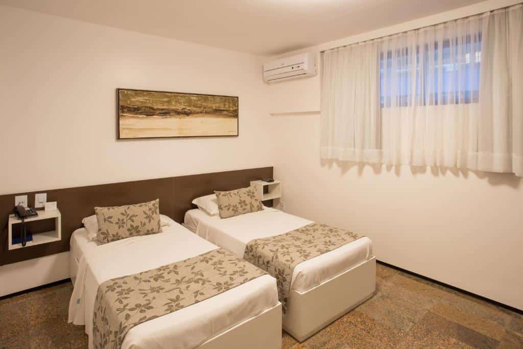 Um quarto de hotel com duas camas de solteiro, colchas florais, uma pintura na parede, ar-condicionado e uma janela com cortinas brancas.