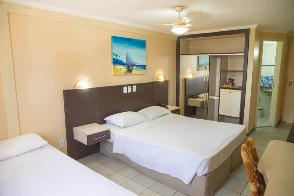 Quarto de hotel com uma cama de casal, uma cama de solteiro, mesinhas de cabeceira, armário espelhado e um banheiro ao fundo.