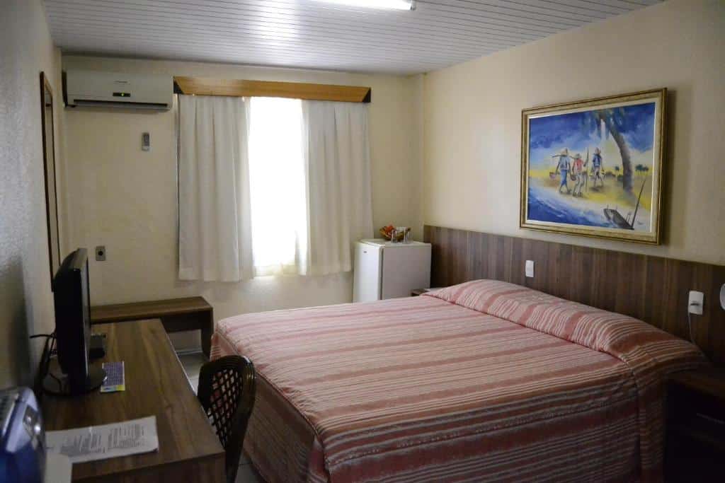 Quarto de hotel com cama de casal, escrivaninha, cadeira, TV, frigobar, ar-condicionado e janela com cortinas fechadas.