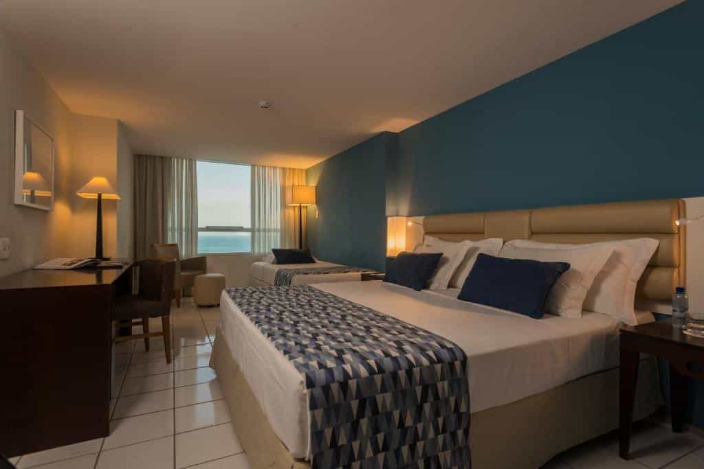 Quarto de hotel com uma cama grande, uma escrivaninha, cadeiras, luminárias, uma janela com vista para o mar e uma parede com detalhes em azul.