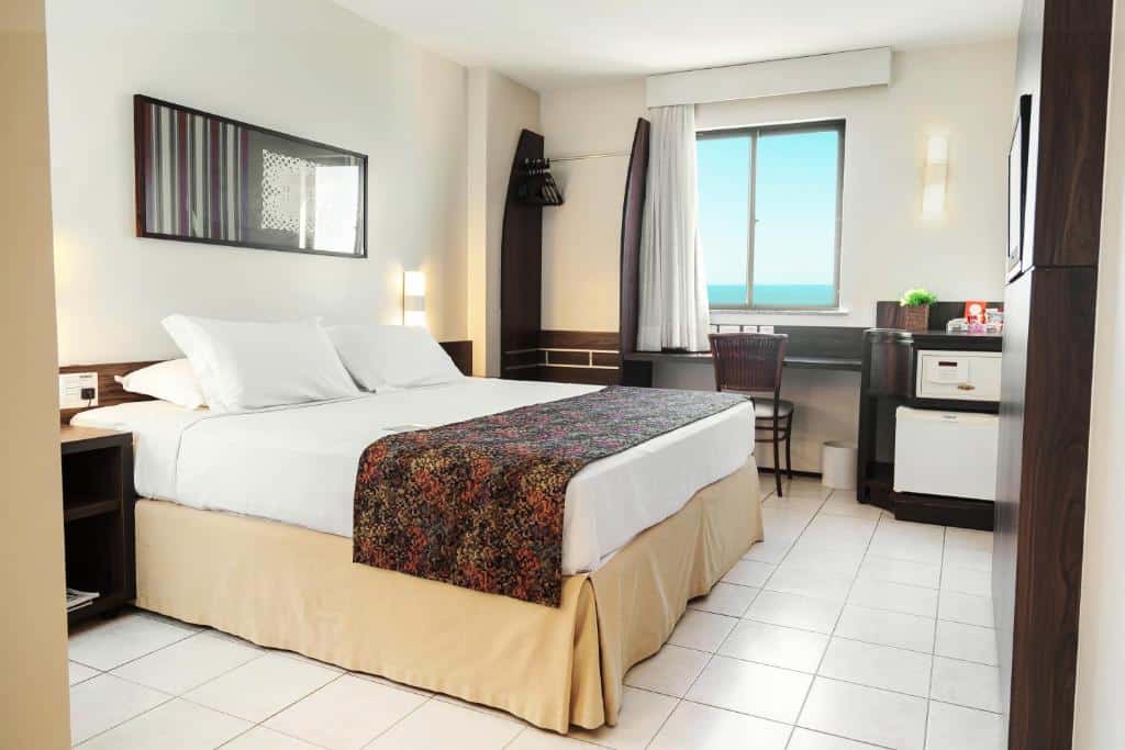 Um quarto de hotel com uma cama de casal, escrivaninha, cadeira, frigobar, espelho de parede e uma janela com vista para o mar.