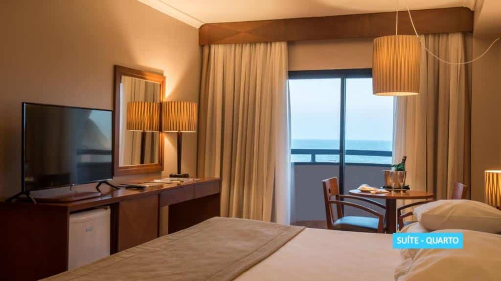 Quarto de hotel com uma cama, escrivaninha, TV, abajur, mesa com duas cadeiras e uma janela com vista para o mar.