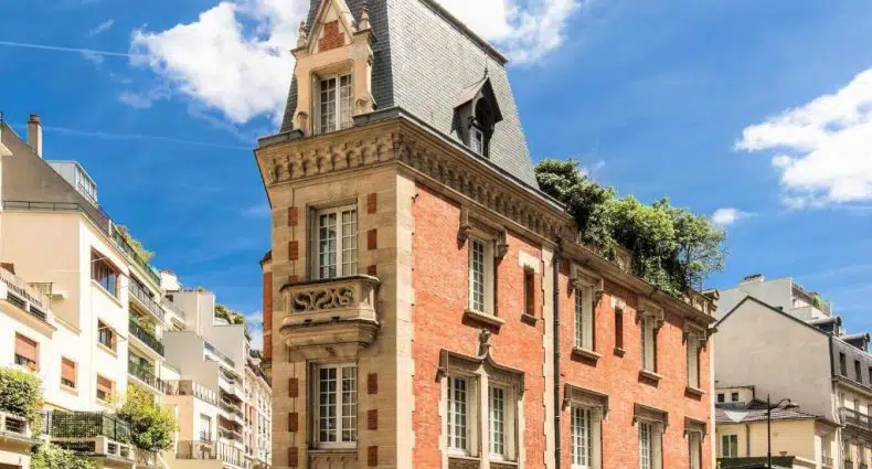 Um prédio estreito de tijolos vermelhos com um telhado cinza íngreme e detalhes ornamentados fica em uma rua da cidade sob um céu azul. Esse é o Sourire Boutique Hôtel Particulier, um dos hotéis boutique em Paris..