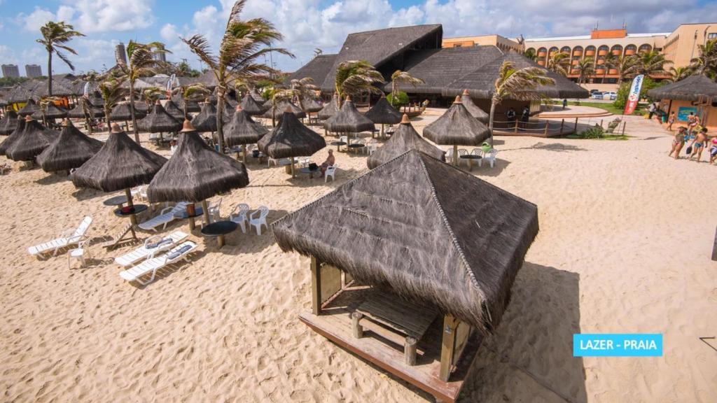 Cabanas com telhado de palha e espreguiçadeiras estão instaladas em uma praia de areia perto de um resort com palmeiras e um céu parcialmente nublado.