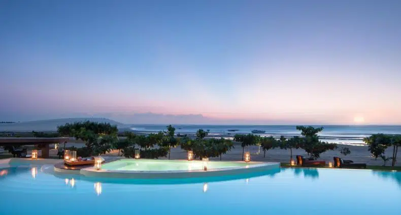 Piscina de borda infinita com vista para o oceano ao pôr do sol, com palmeiras e lanternas ao redor da área da piscina.
