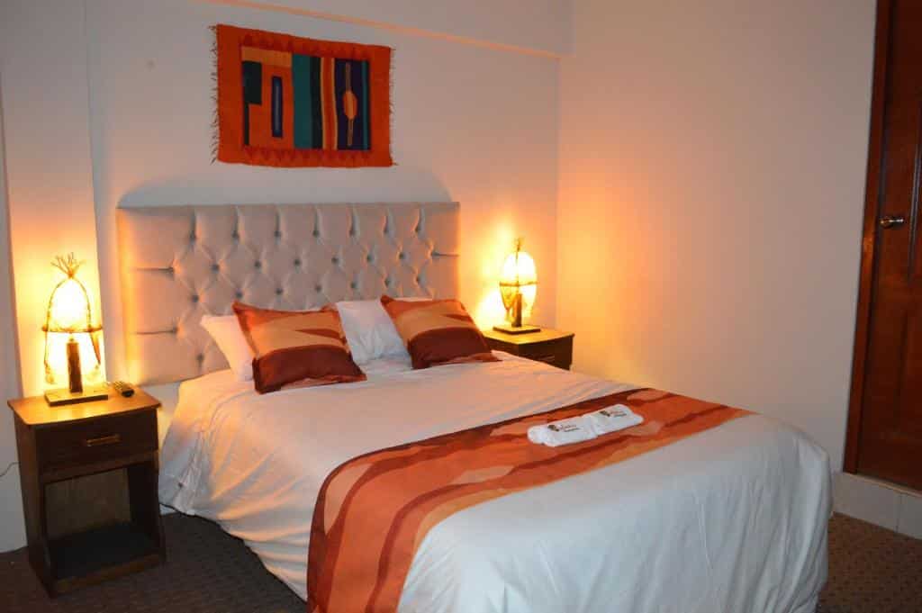 Quarto de casal do Andino Hotel barato com uma cama de casal à esquerda com uma cômoda de cada lado da cama, há um abajur em cada uma.