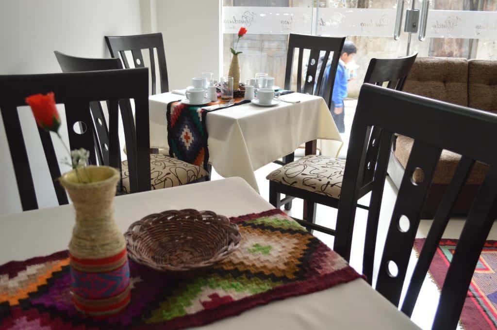 refeitório do Andino Hotel barato com duas mesas de madeira quadradas, cada uma com quatro cadeiras. Em cima da mesa há um enfeito de tecido temático decorando, além de um vaso com uma flor sintética e xícaras e copos.