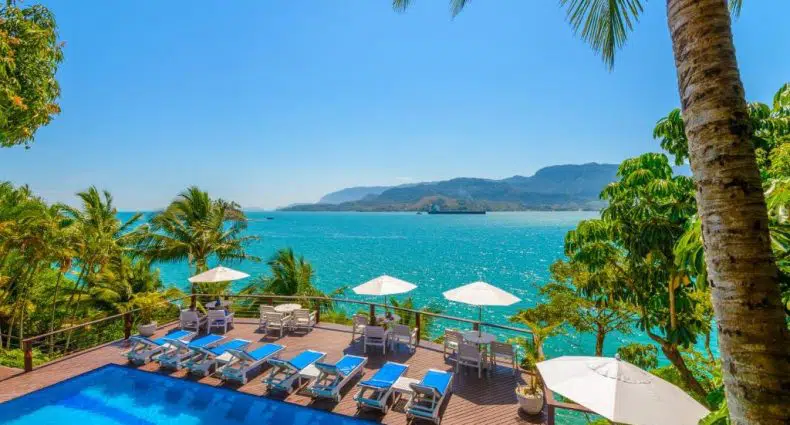 Uma área de piscina com espreguiçadeiras e guarda-sóis azuis tem vista para um mar azul-turquesa, cercada por palmeiras e uma paisagem montanhosa no Barra do Piuva Porto Hotel, para representar onde passar o Réveillon em São Paulo