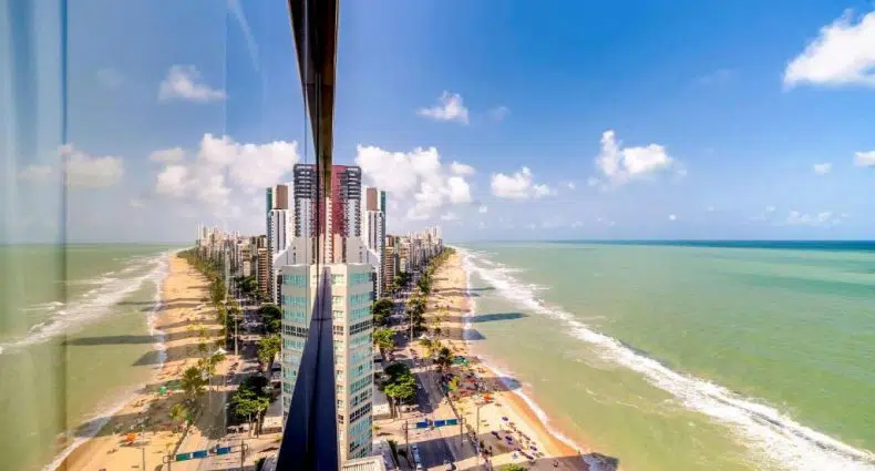 Prédios altos e uma praia são refletidos em uma janela de vidro, criando uma visão dividida simétrica do litoral.