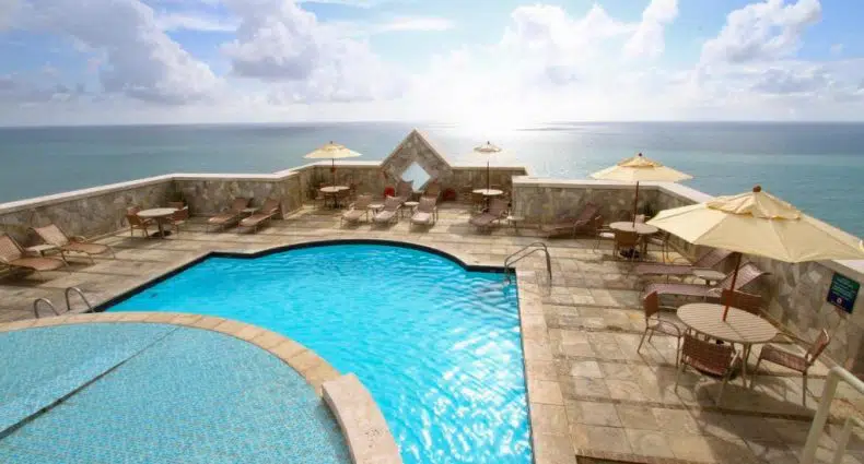 Piscina na cobertura com espreguiçadeiras, mesas e guarda-sóis com vista para o oceano sob um céu parcialmente nublado.