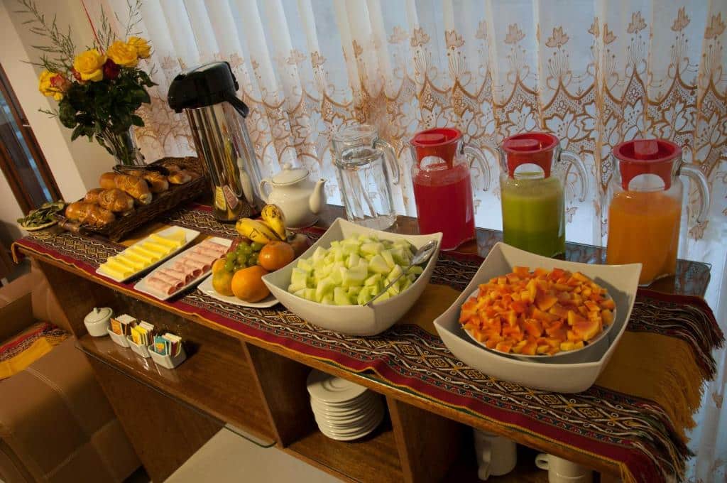 mesa de café da manhã do Picos House com duas tigelas de frutas, quatro jarras de sucos e água, uma garrafa térmica com café, dois pratos de presunto e queijo e uma cesta de pães