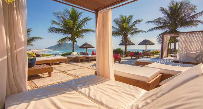 Vista de uma cabana sombreada com vista para um pátio com espreguiçadeiras, palmeiras e o oceano ao fundo.