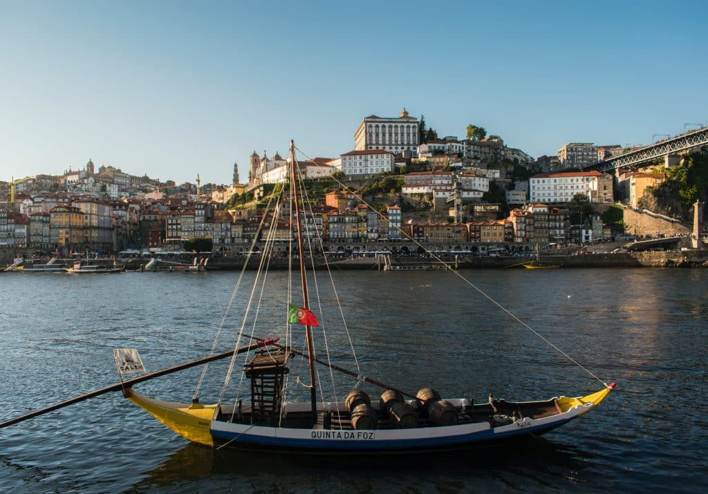 Barco no Cais da Ribeira dentro do rio Douro durante o dia e ao fundo várias casinhas.