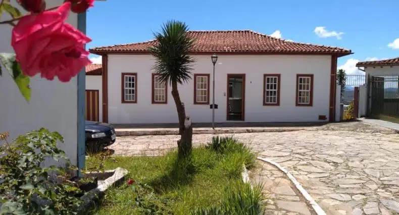Casa branca de um andar com telhado marrom, passarela de pedra, grama verde, rosas e uma árvore em primeiro plano em um dia ensolarado.