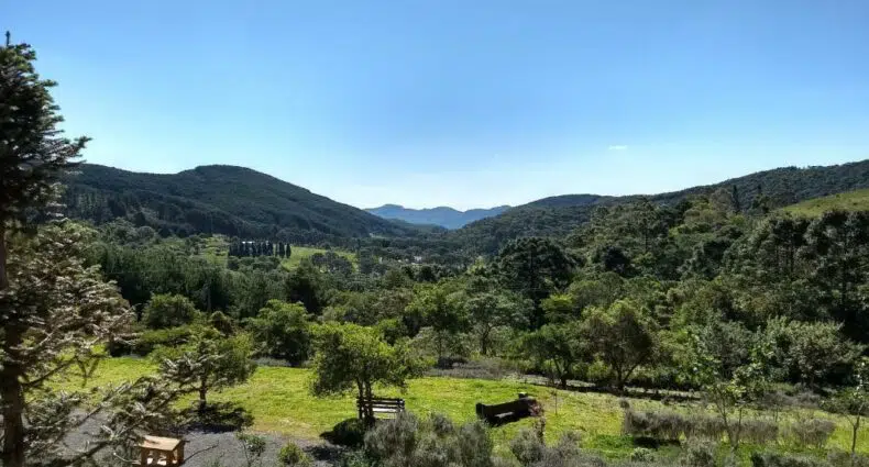 Vista da Pousada Pinho Bravo, uma das pousadas em Camanducaia, de um vale verde, coberto de árvores, com colinas à distância, sob um céu azul claro.