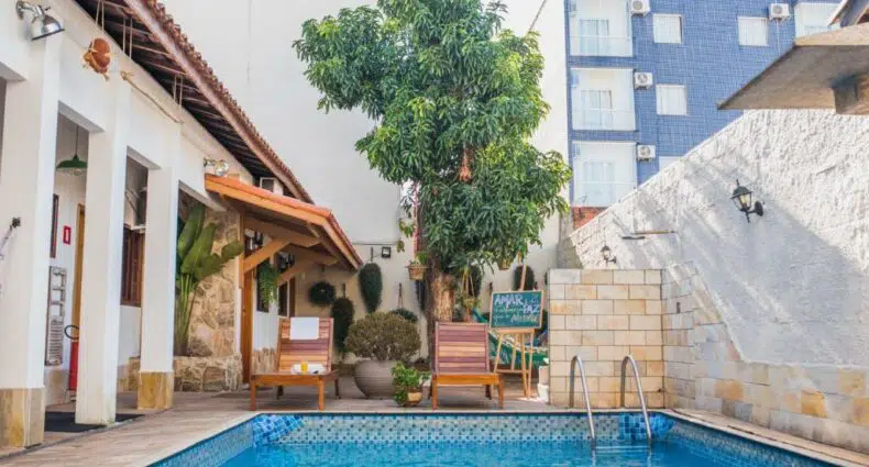 Área de piscina ao ar livre na Pousada Rogai com duas espreguiçadeiras de madeira, uma pequena árvore, uma placa de lousa e prédios ao redor ao fundo.