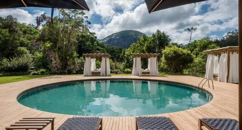 Piscina circular ao ar livre com espreguiçadeiras, cabanas e vista para a montanha, cercada por árvores sob um céu parcialmente nublado.