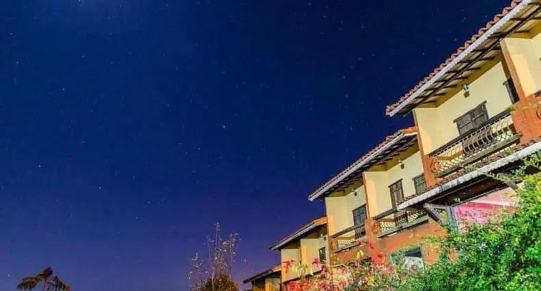 Um céu noturno com estrelas acima de um edifício de vários andares com varandas e algumas árvores em primeiro plano.