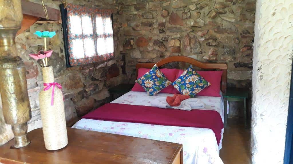 quarto da Pousada Casa do Arco com uma cama de casal no meio do quarto e uma cômoda de madeira em frente. As paredes são todas de pedras expostas