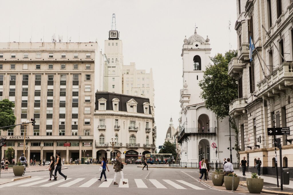 Foto de uma faixa de pedestres com pessoas atravessando a rua em Buenos Aires, Argentina. Os prédios ao redor são antigos, mas conservados. Há também poucas árvores ao redor. A imagem tem tons pálidos e o céu está completamente branco. - Foto: Sasha Stories via Unsplash