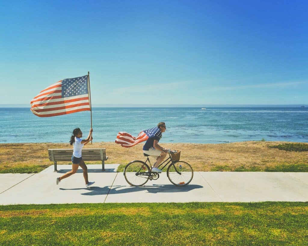 Homem andando de bicicleta com uma bandeira dos EUA amarrada aos ombros, como uma capa de super-herói, ao centro da imagem, seguido por uma moça de rabo de cavalo, camisa branca e shorts jeans, também carregando uma bandeira dos EUA, porém com um mastro, e ao fundo vê-se o mar. - Foto: frank mckenna via Unsplash