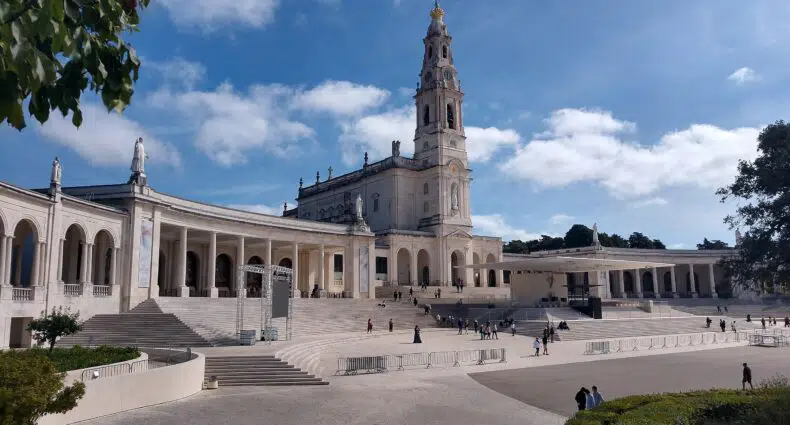 Grande basílica branca com um alto campanário e uma colunata em arco sob um céu azul, com pessoas caminhando ao redor, trata-se do Santuário de Fátima, para representar hotéis em Fátima.