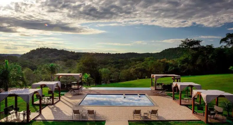 Piscina externa cercada por espreguiçadeiras e cabanas, com vista para colinas gramadas e árvores sob um céu nublado.
