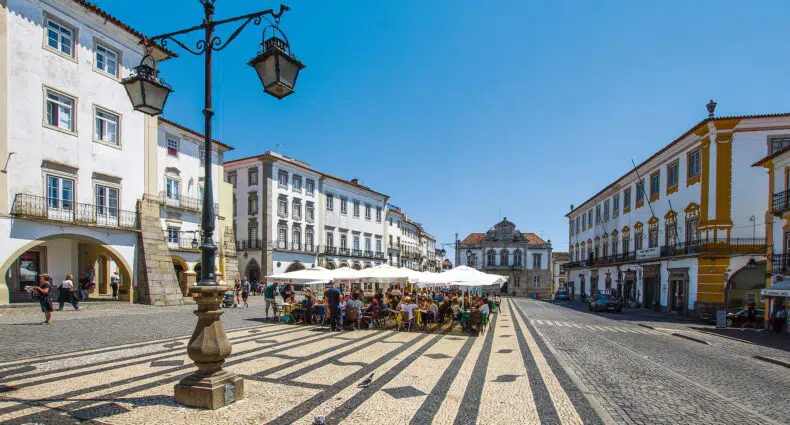 Café ao ar livre na Praça do Giraldo em Évora com guarda-chuvas brancos em uma praça de paralelepípedos, cercado por edifícios históricos brancos sob um céu azul claro, para ilustrar o post de hotéis em Évora.