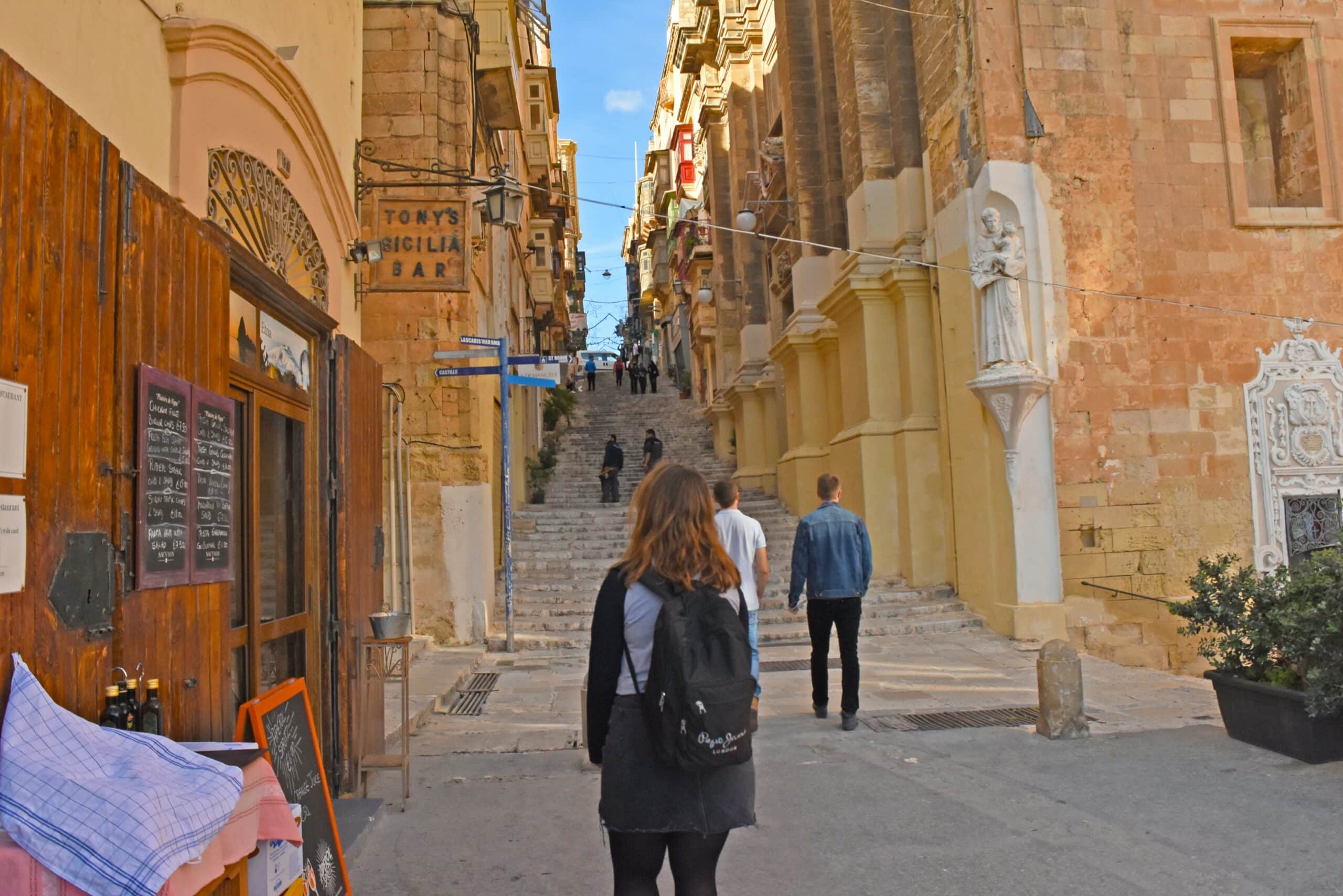 Uma rua estreita, com escadaria e rodeada por construções antigas em Malta, na Europa. Também há uma mulher, de cabelo avermelhado, com mochila nas costas, e dois homens indo em direção aos degraus
