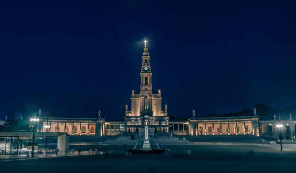 Santuário de Fátima, em Portugal, iluminado à noite com uma cruz acesa no topo da torre central e uma praça vazia em frente.