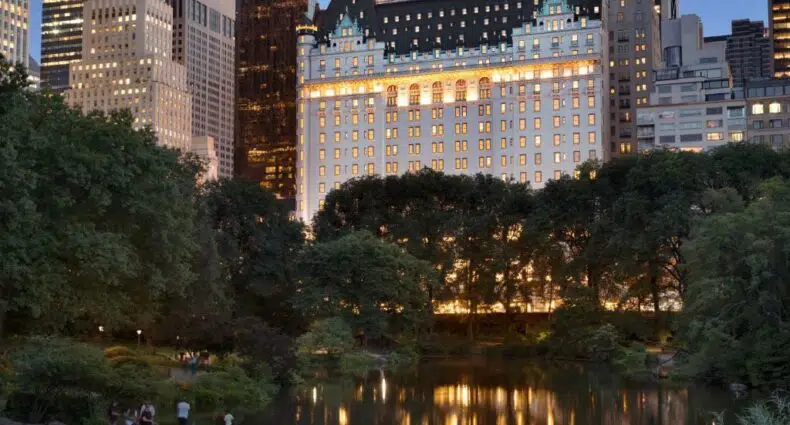 Um prédio grande e bem iluminado, trata-se do The Plaza Central Park, atrás de árvores e de um lago ao anoitecer em uma cidade com prédios altos, para representar hotéis de luxo em Nova York.