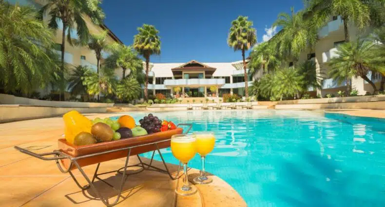 Uma cena à beira da piscina com uma bandeja de frutas variadas e dois copos de suco de laranja no deque, cercada por palmeiras.