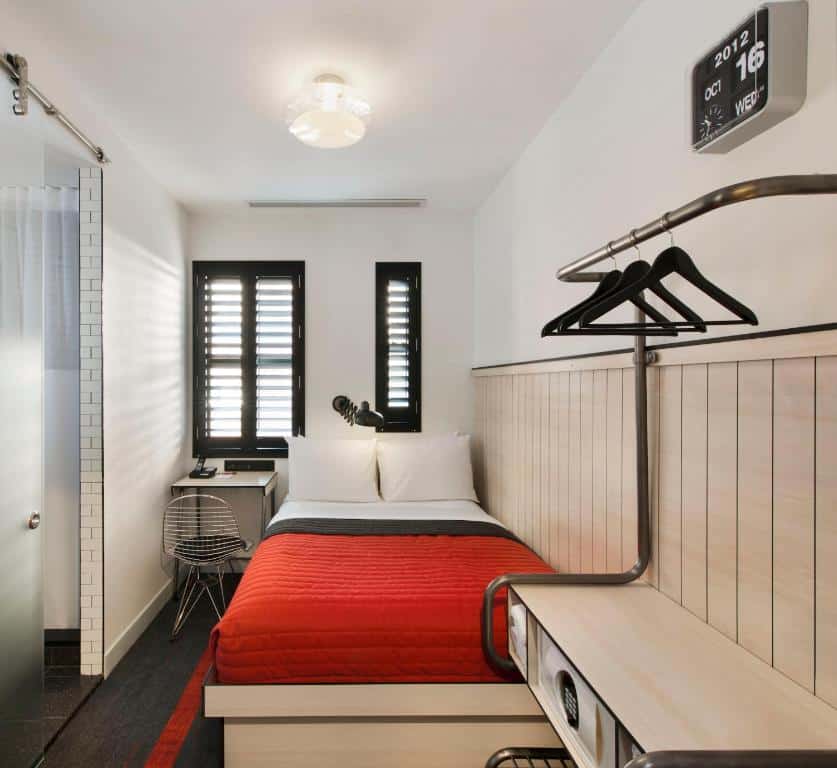 quarto individual do Pod 39 com uma cama de casal pequena, com acolchoado vermelho, uma pequena mesa de canto no lado esquerdo da cama e uma porta de vidro de correr no lado esquerdo, apontando para o banheiro privativo.