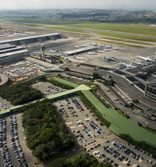 Vista aérea do Aeroporto de Guarulhos, sendo possível enxergar pistas com aviões, uma área com corredores para embarque e desembarque, carros estacionamentos ao ar livre e algumas árvores por perto