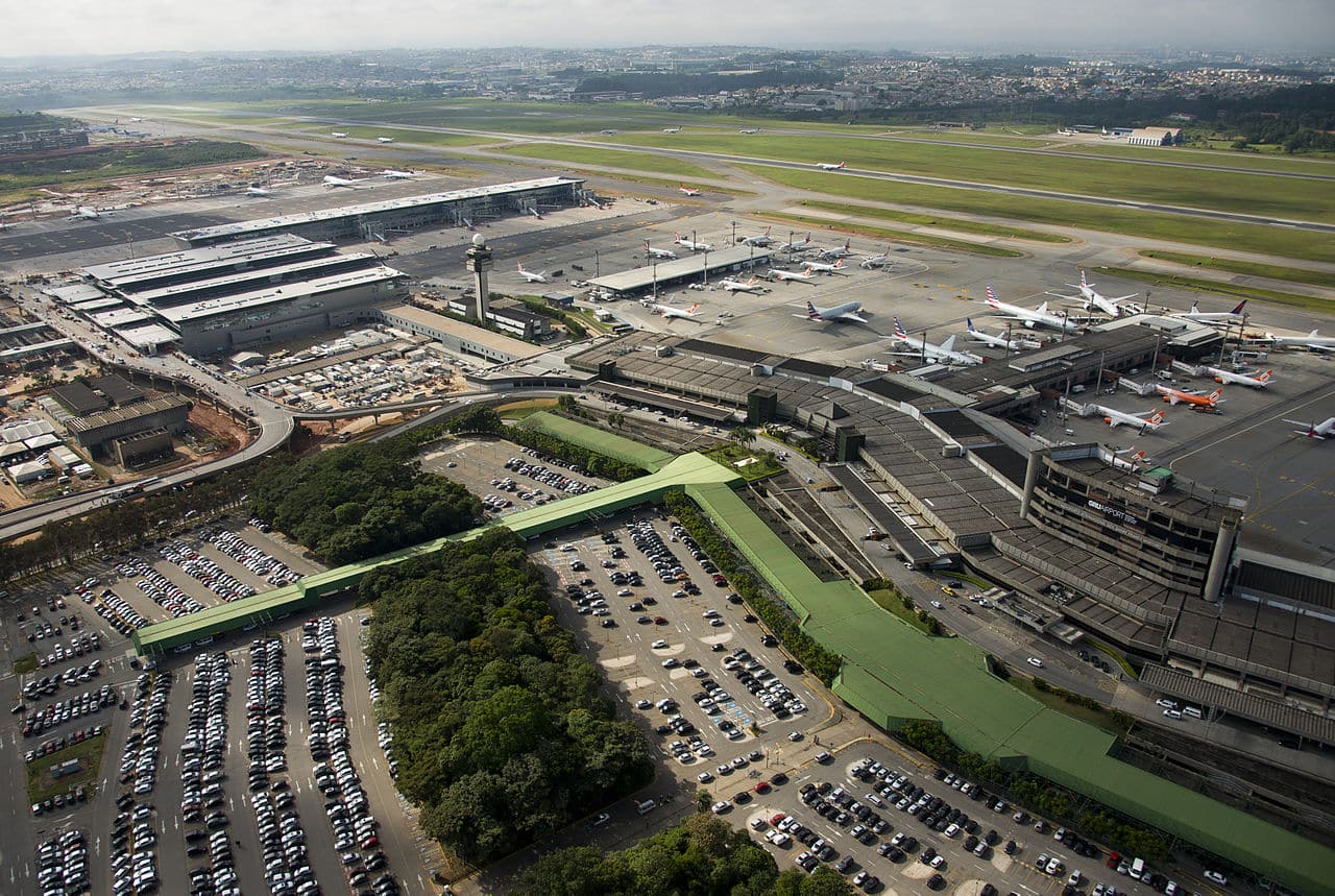 Vista aérea do Aeroporto de Guarulhos, sendo possível enxergar pistas com aviões, uma área com corredores para embarque e desembarque, carros estacionamentos ao ar livre e algumas árvores por perto