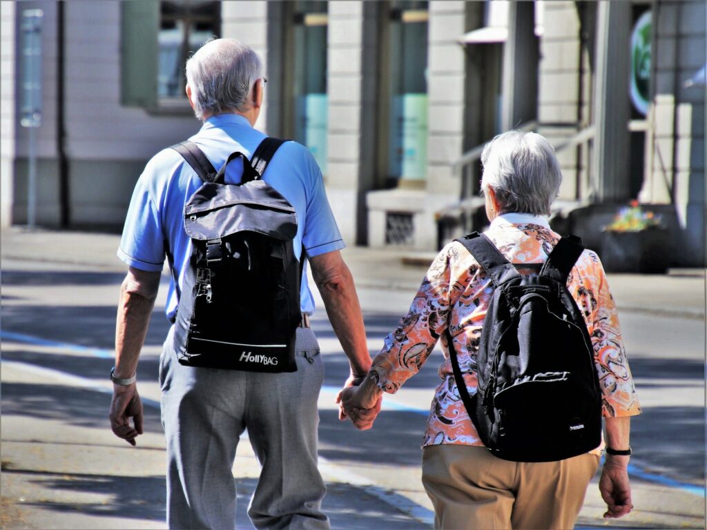 Casal idoso passeando de mãos dadas durante o dia. Eles estão de costas para a foto e usam mochilas. Foto: Pasja 1000 via Pixabay