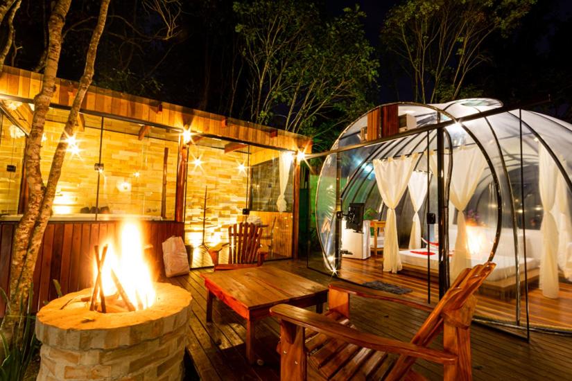 Fogueira acesa em uma noite no Chalé Luiz, com uma tenda transparente  iluminada com cama de casal (estilo japonesa) e várias cortinas dentro. Há também árvores e natureza em volta